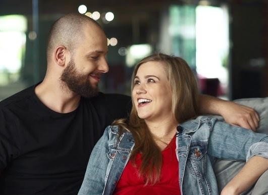 Maciej Pela w swobodnej, zamyślonej pozie w casualowym stroju, na tle rozmytego wnętrza.