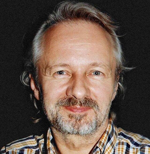 Krzysztof Stroiński na planie filmowym, w kostiumie, z widocznym sprzętem filmowym w tle.