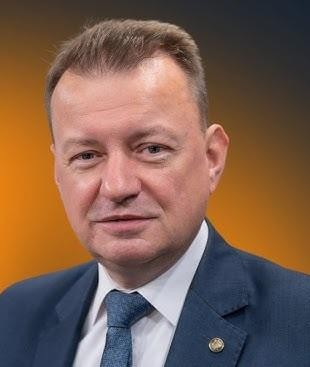 Mariusz Błaszczak przemawiający z mównicy, z gestykulacją, na tle rozmytego tłumu słuchaczy lub innych polityków.