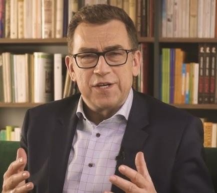 Maciej Orłoś podczas wystąpienia publicznego, z pasją gestykulujący, w profesjonalnym stroju, na tle sceny.