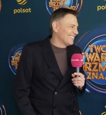 Krzysztof Ibisz w studiu telewizyjnym podczas prezentowania programu, dynamicznie gestykulujący, co podkreśla jego energię i wszechstronność.
